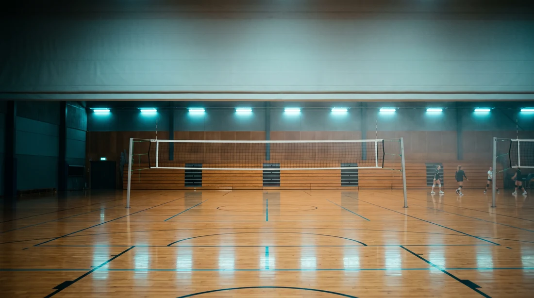 Eine Sporthalle am Abend: das Netz gespannt, der Hallenboden mit den Spielfeldlinien spiegelt die Deckenlichter, hinten die Bank.